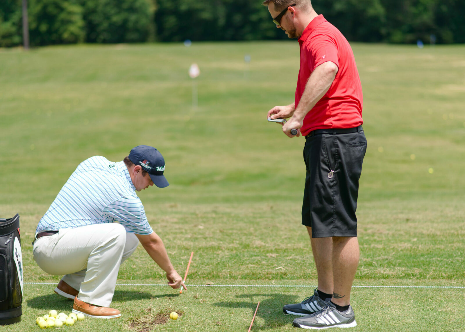 Two men on a golf course at Williamsburg National; one in a striped shirt and cap kneels, adjusting a golf ball and stick. The other in a red shirt and sunglasses stands, holding a device, observing. They are on a grassy field with scattered golf balls and trees in the background.